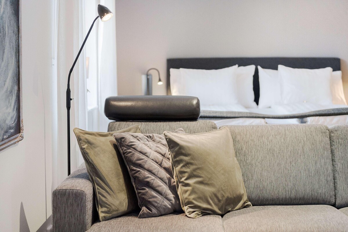 A modern hotel room with a grey sofa, two pillows, a floor lamp, and a bed with white linens.