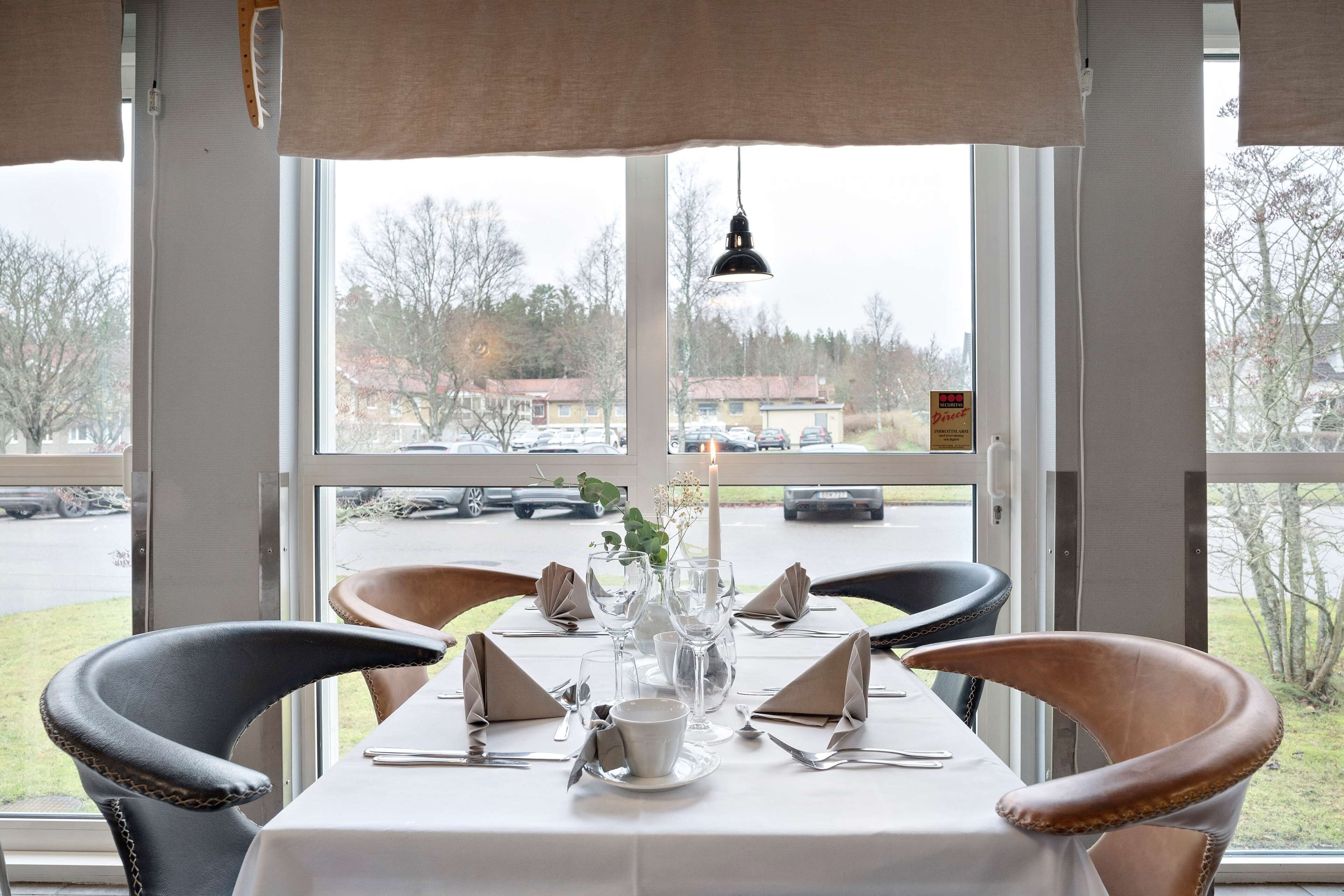 Une table à manger dressée pour deux personnes, agrémentée d’un vase de fleurs et d’une bougie, avec vue sur un parking et des immeubles, le tout visible à travers de grandes fenêtres.