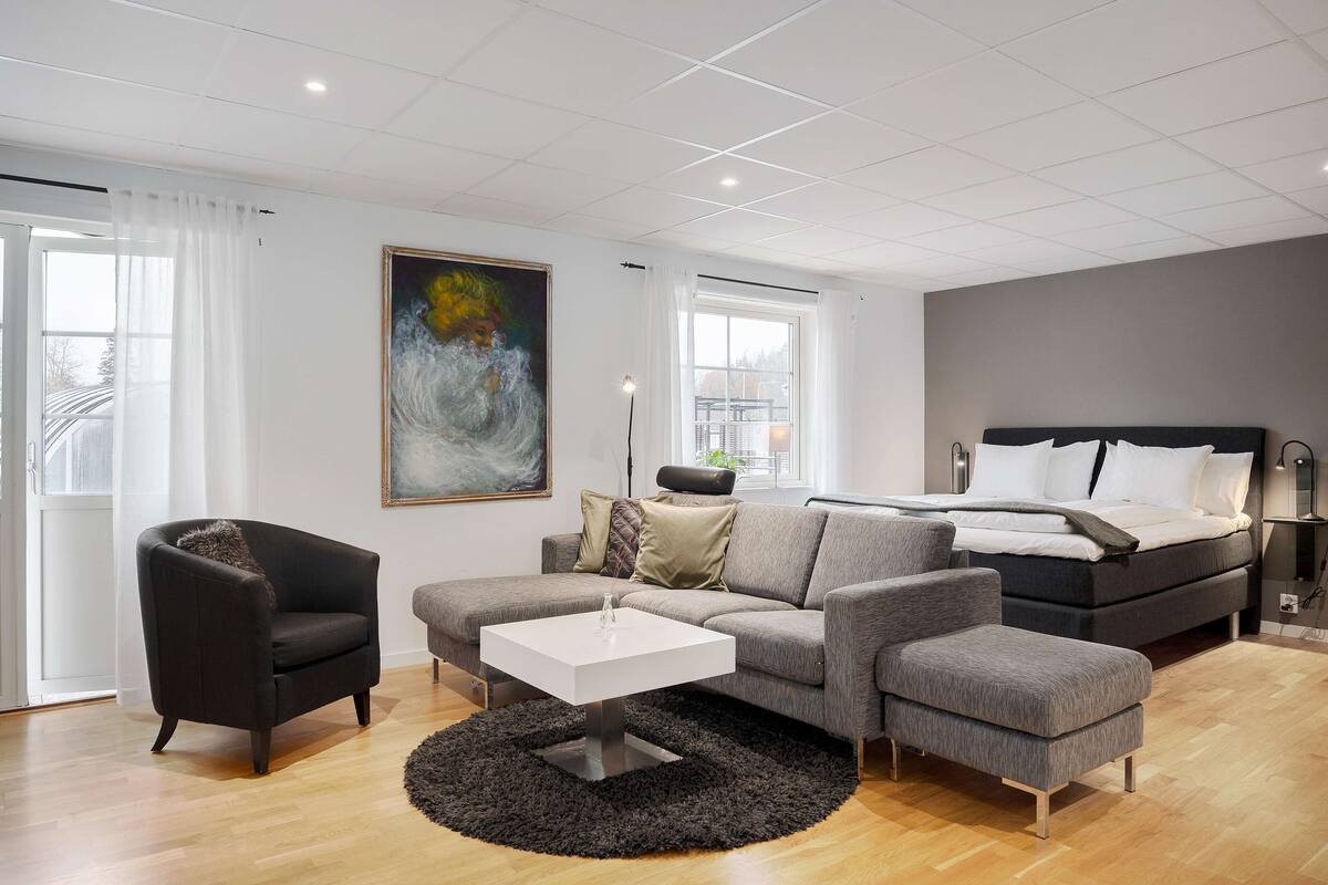 A modern living room with a grey sofa, a black armchair, a white coffee table, and a grey ottoman. There is a large painting on the wall, a bed with white bedding, and a window with curtains.