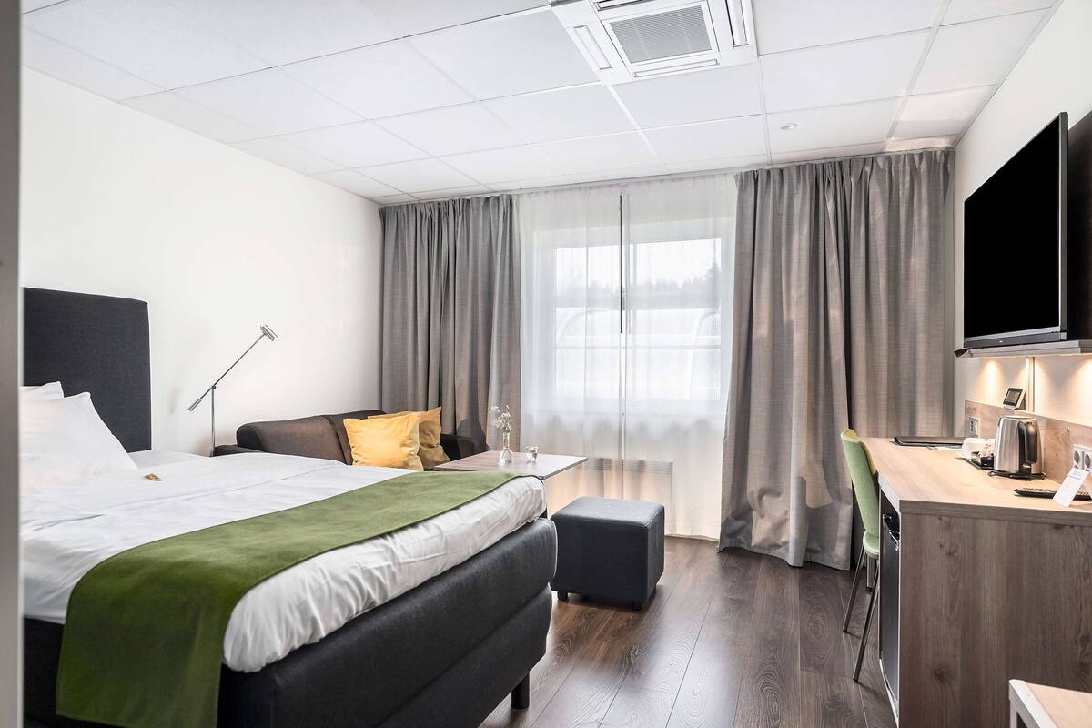 A modern hotel room with a large bed, a desk, a flat-screen TV, and a window with curtains.