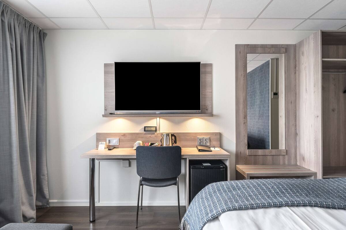 A modern hotel room with a bed, a desk with a chair, a television, a small kitchenette, and a wardrobe.
