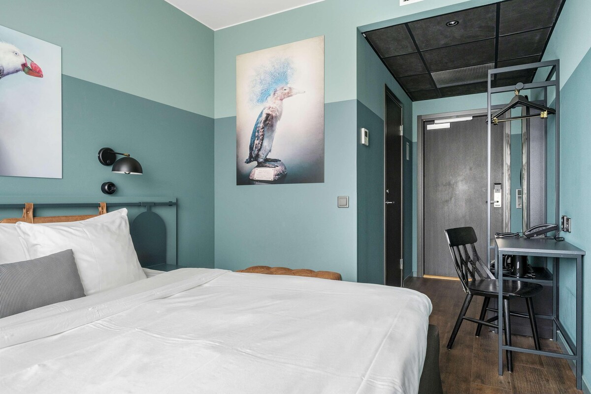 A modern hotel room with a bed, a desk, and a chair. There is a painting on the wall and a lamp on the nightstand.
