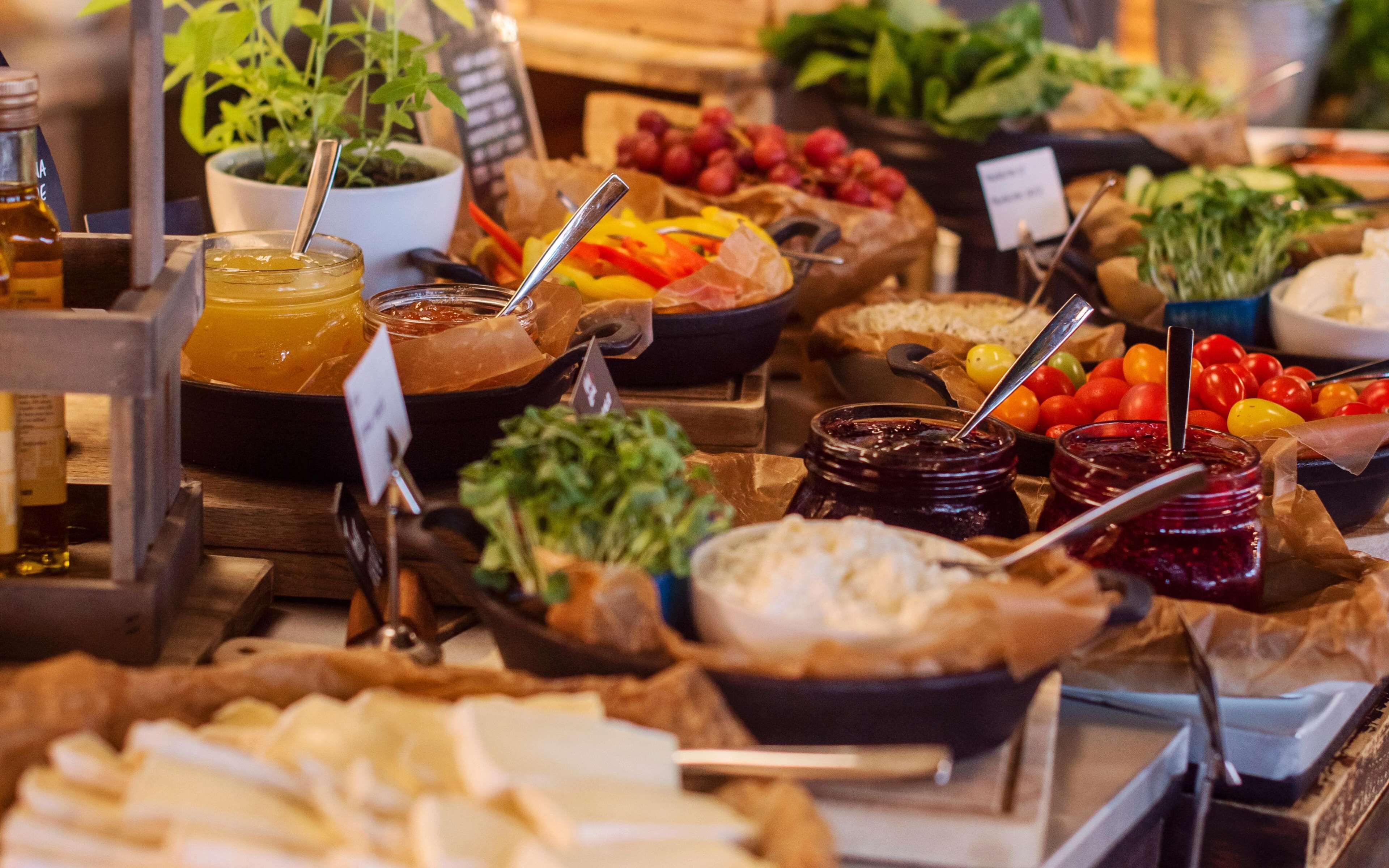 Variedad de alimentos y condimentos expuestos sobre una mesa, incluyendo frascos de mermelada, tazones de verduras y un recipiente con líquido amarillo.