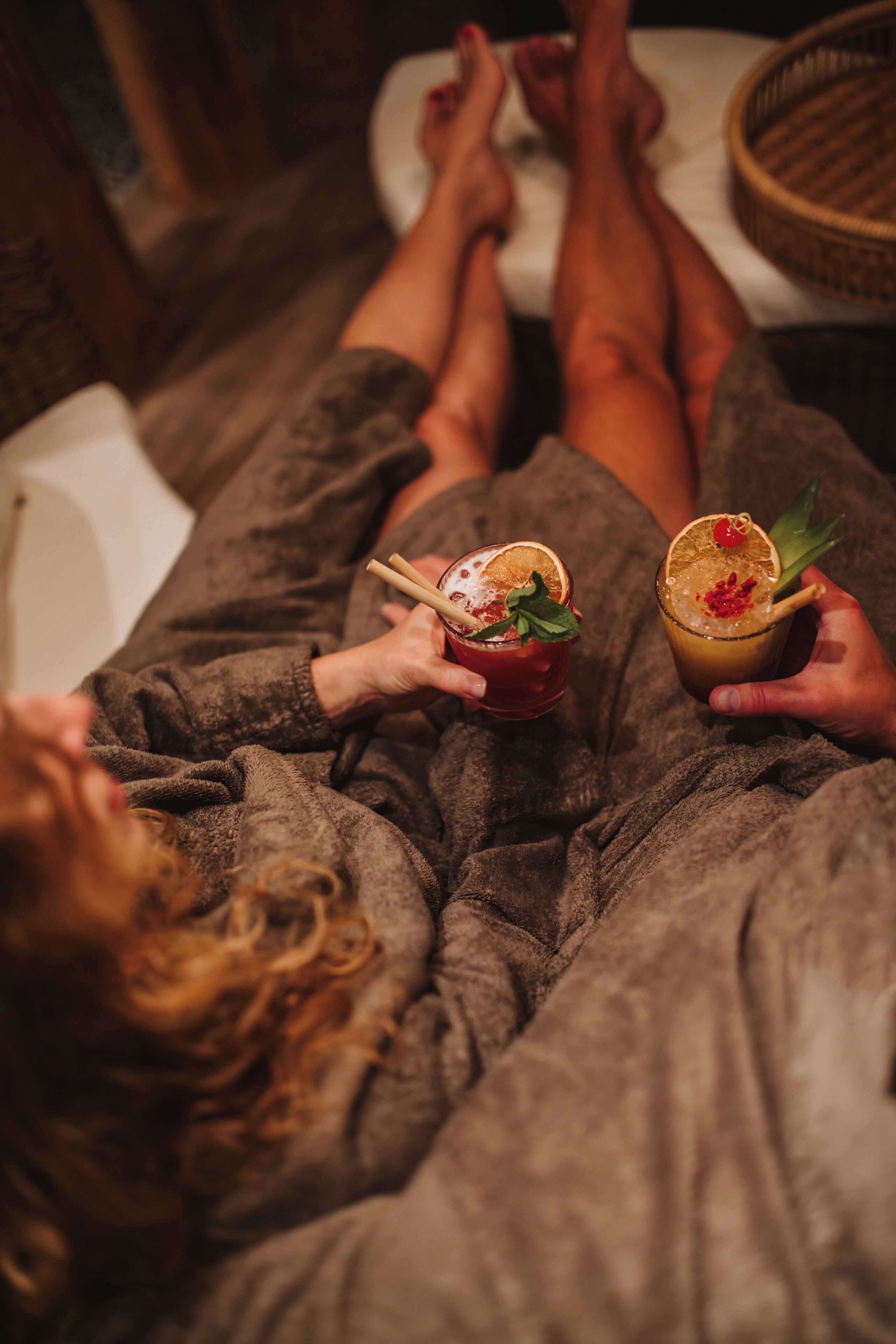 A person lying on a bed in a robe, holding a drink with a garnish.