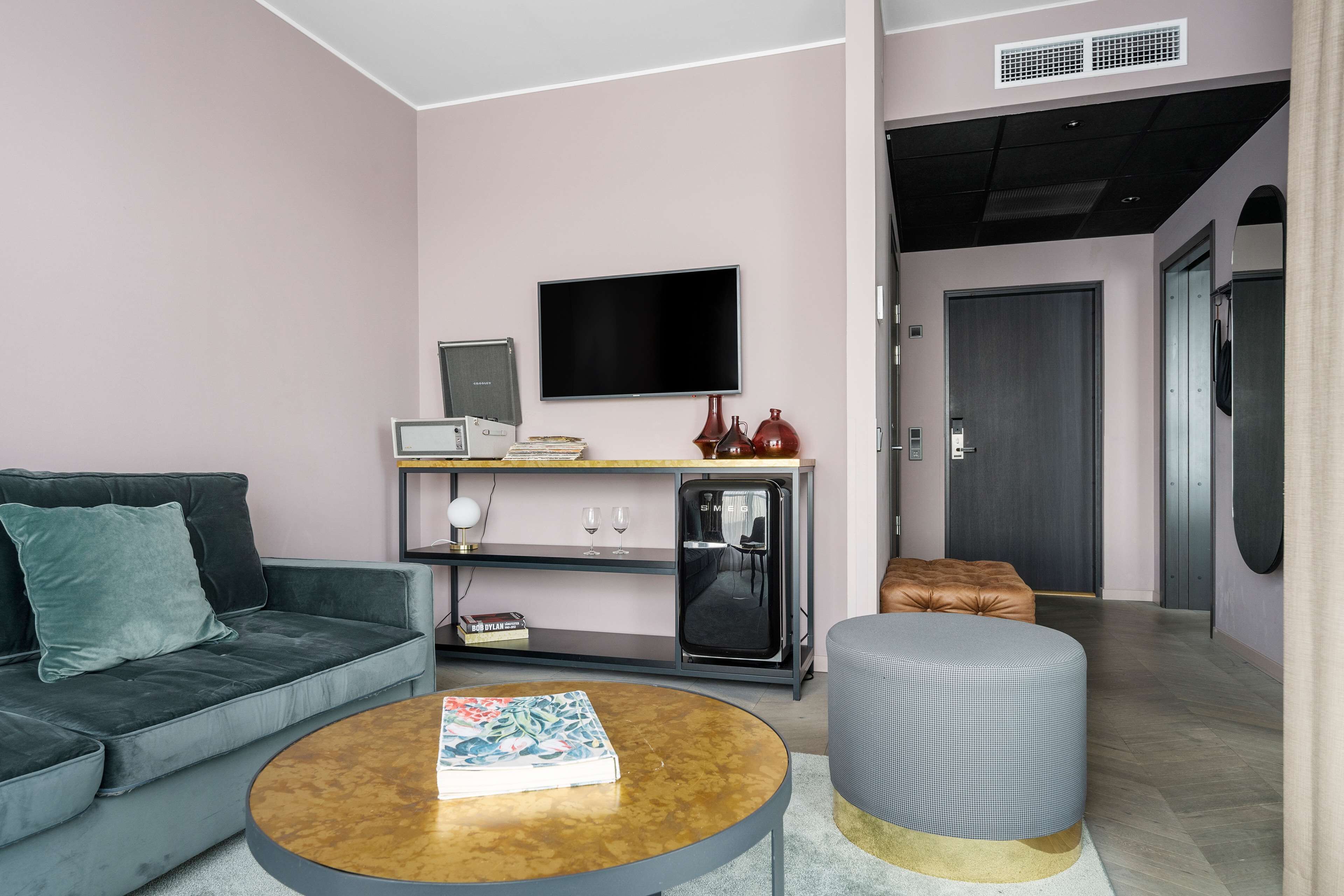 Una sala moderna con un sofá gris, una mesa redonda de centro, un televisor de pantalla plana, un refrigerador pequeño y una puerta que da a otra habitación.