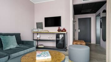 Una sala moderna con un sofá gris, una mesa redonda de centro, un televisor de pantalla plana, un refrigerador pequeño y una puerta que da a otra habitación.