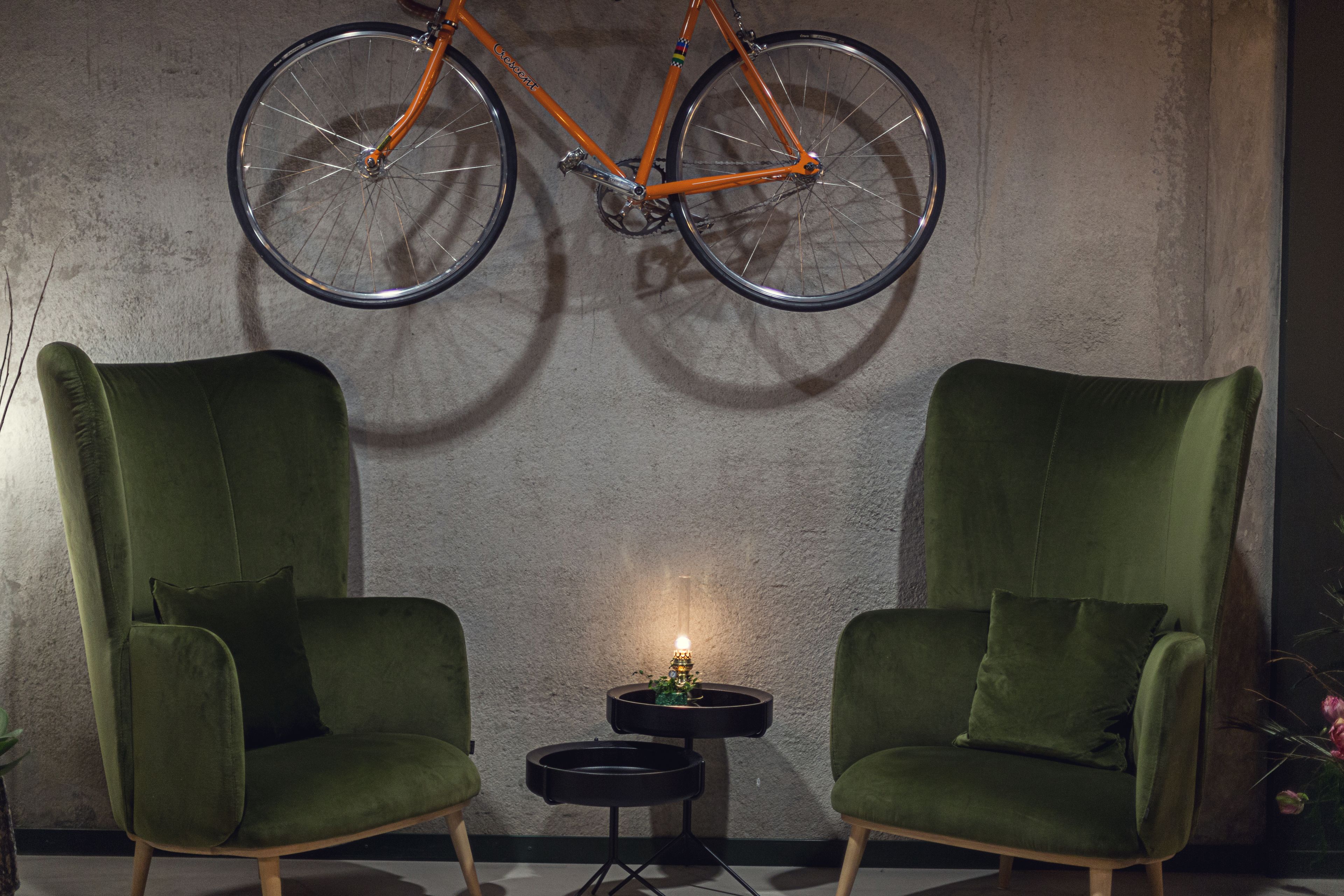 A room with two green armchairs, a small round table with a candle, and an orange bicycle hanging from the ceiling.