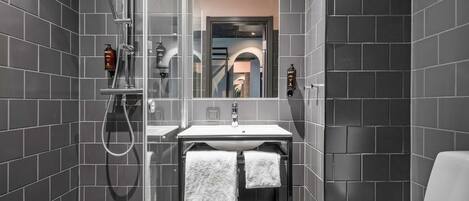 A modern bathroom with dark tiled walls, a glass shower enclosure, a white sink, and two towels hanging on a rack.