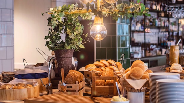 A buffet setup with various breads, a potted plant, and a bar area in the background. - Best Western Plus Hus 57 (Angelholm)