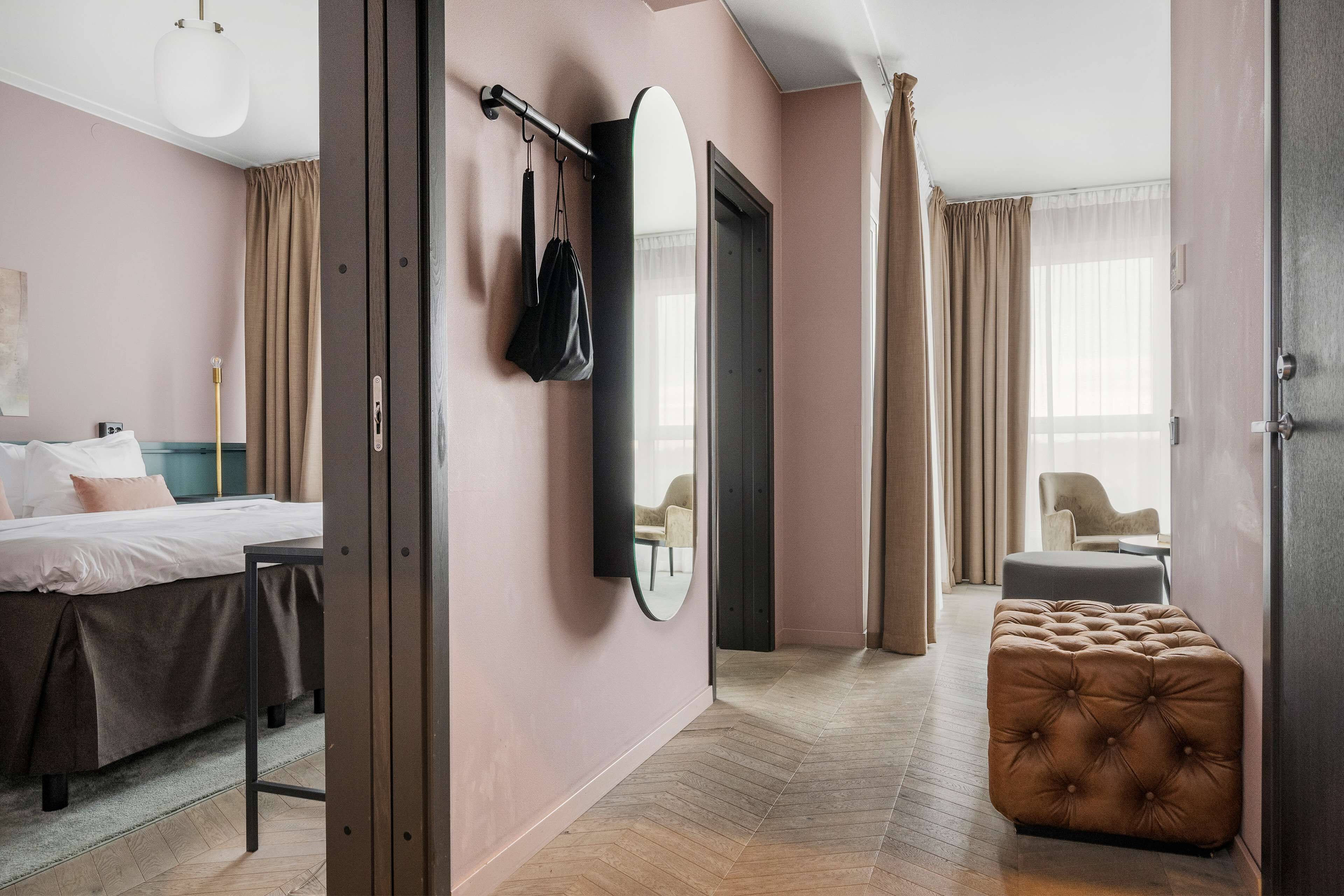 Une chambre d’hôtel moderne comprenant un lit, un coin salon avec un fauteuil, un grand miroir et un pouf capitonné.