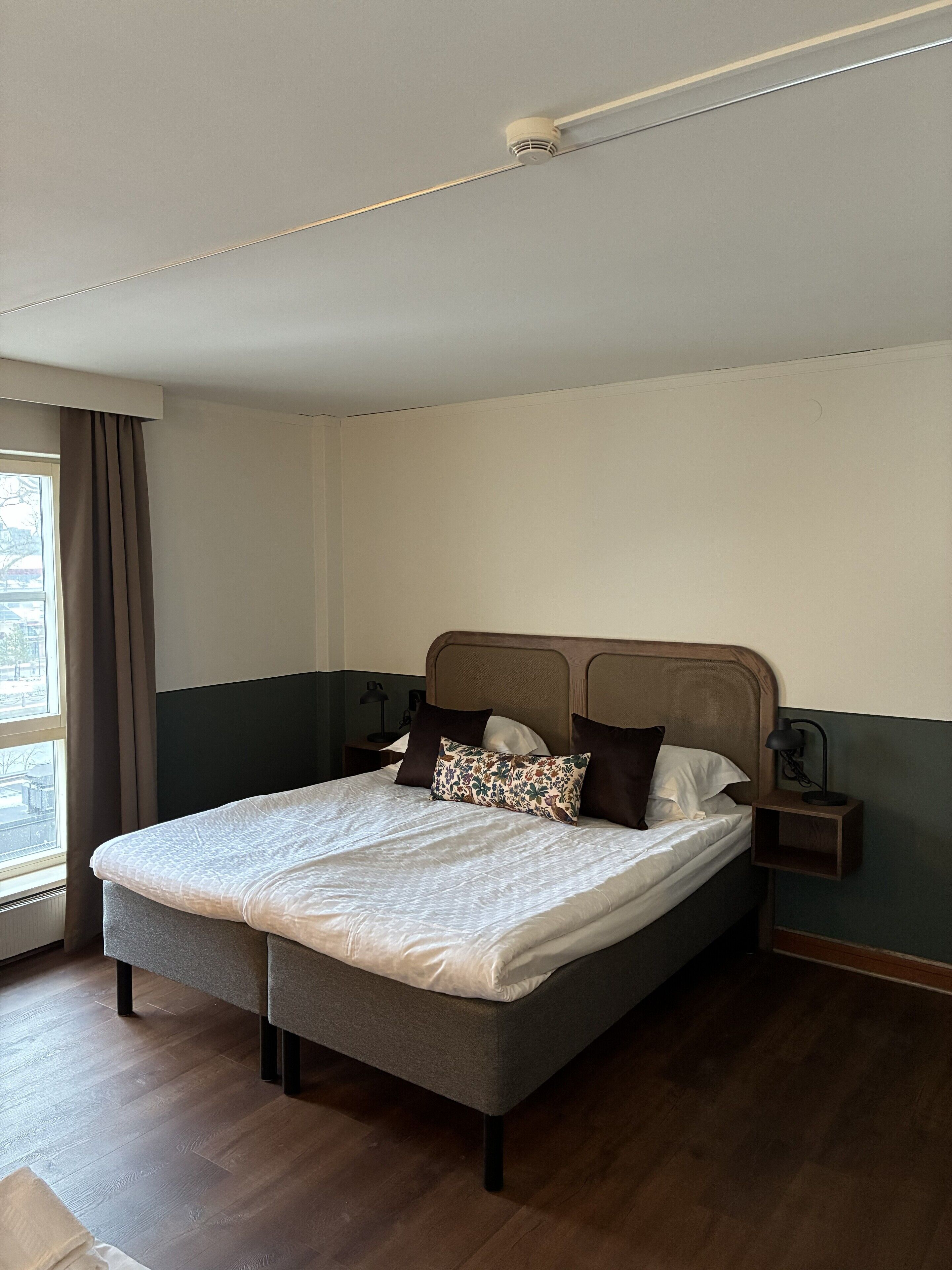 A neatly made bed with a patterned pillow, a bedside table with a lamp, and a window with curtains.