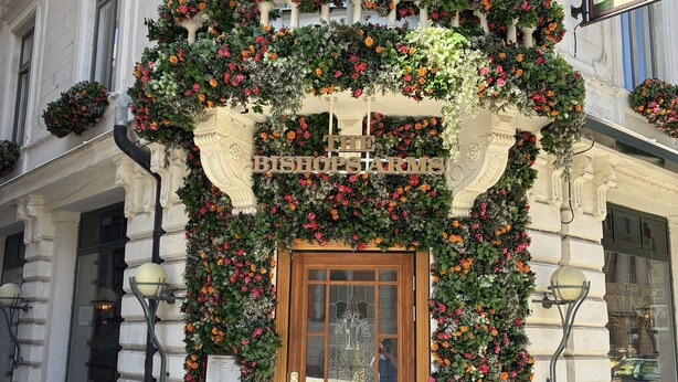 Entrada de hotel con puerta de madera, cartel con tarifas de habitaciones y un arco decorado con flores.