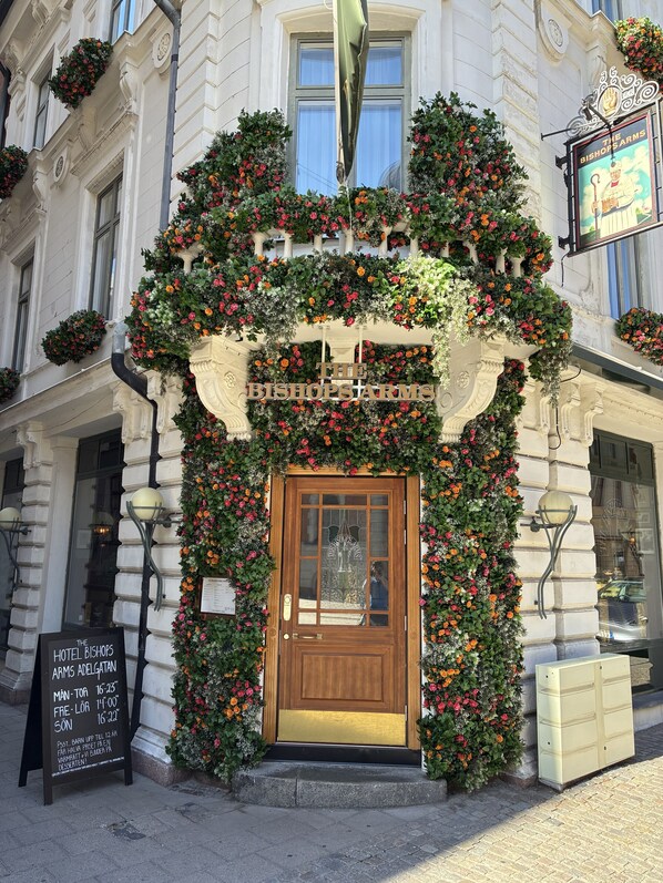 Entrada de hotel com porta de madeira, placa de preços de quartos e arco decorado com flores.