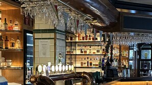 Una zona de bar con taburetes de madera, una barra pulida y estantes llenos de botellas y vasos.