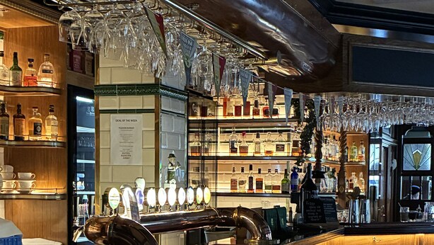 Una zona de bar con taburetes de madera, una barra pulida y estantes llenos de botellas y vasos.