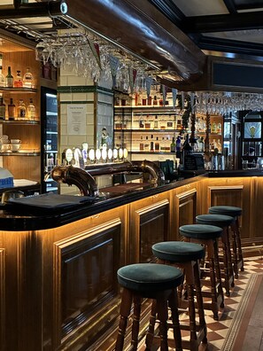 Una zona de bar con taburetes de madera, una barra pulida y estantes llenos de botellas y vasos.