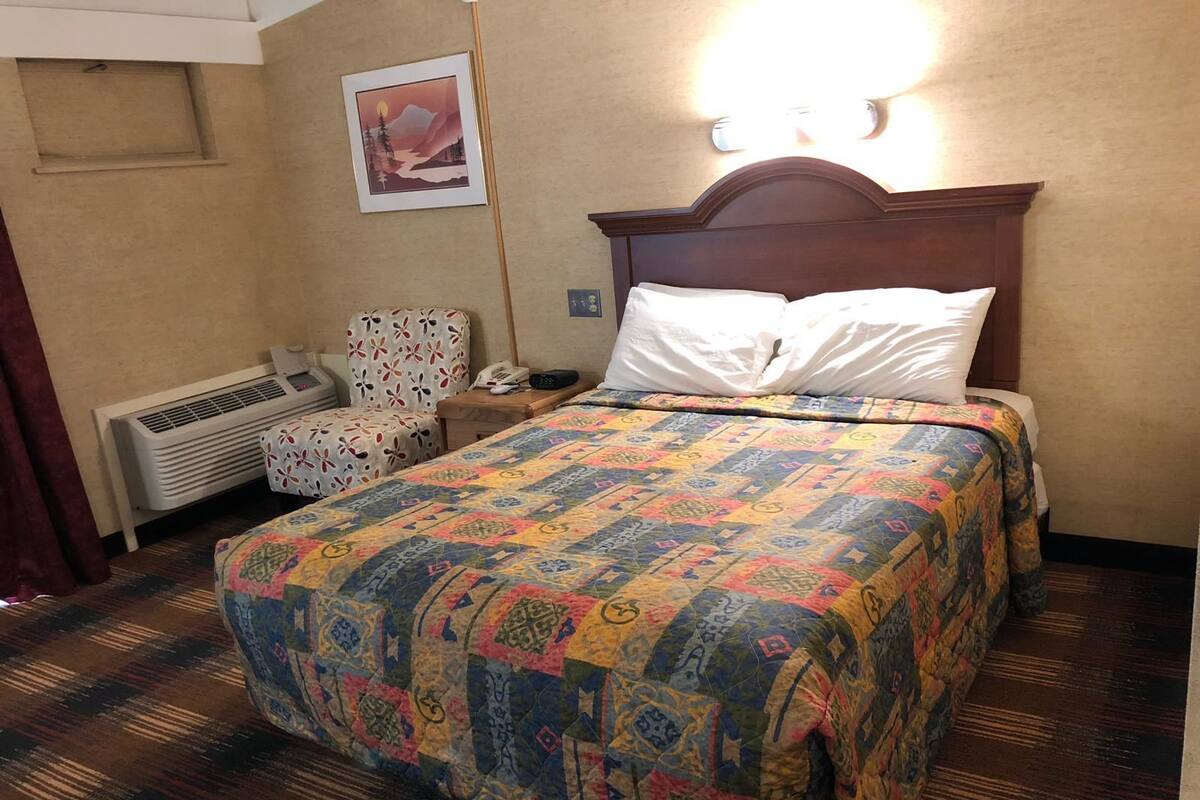 A hotel room with a large bed, a chair, an air conditioning unit, a bedside table with a phone, and a wall-mounted light.