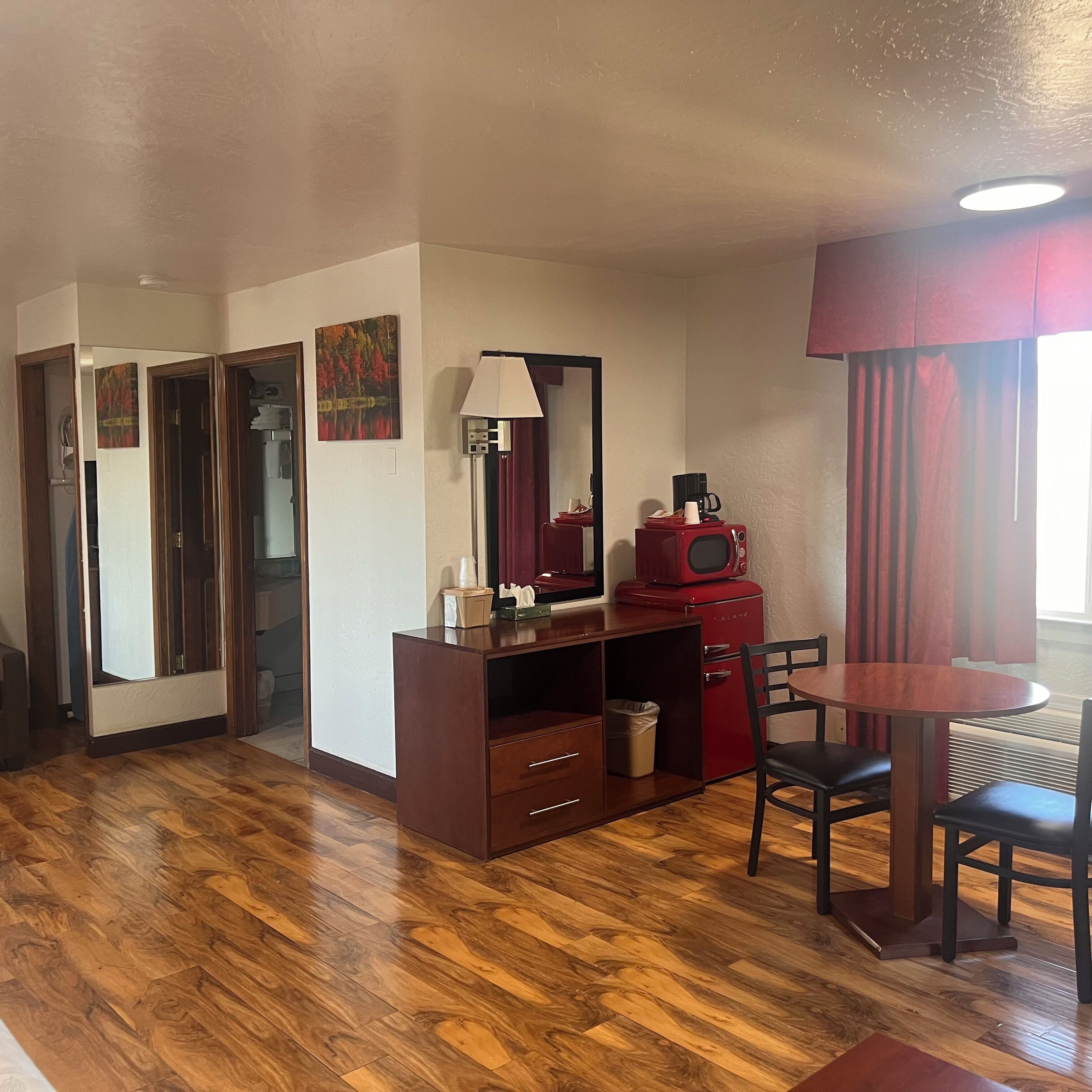 Una habitación compacta con suelo de madera, un escritorio con cajones, un microondas rojo, una mesa con sillas y una ventana con cortinas.
