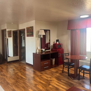 A compact room with a wooden floor, a desk with drawers, a red microwave, a table with chairs, and a window with curtains. - Red Carpet Inn Medford (Medford)