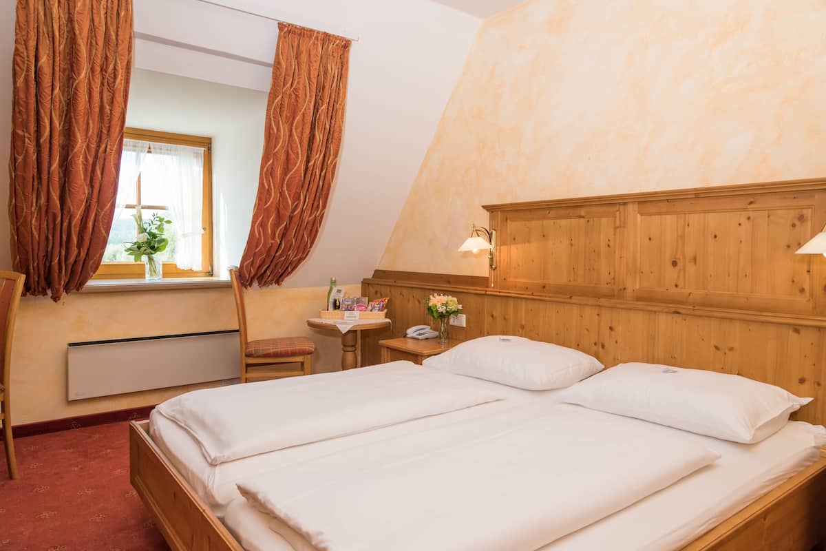 Une chambre à coucher avec une tête de lit en bois, un lit recouvert de draps blancs, une petite table sur laquelle est posé un vase de fleurs et une fenêtre avec des rideaux.