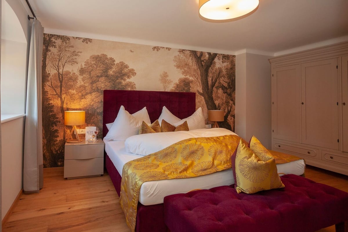 Une chambre à coucher avec un grand lit, deux tables de chevet, une commode, une armoire et un mur décoré d’une fresque représentant un arbre.