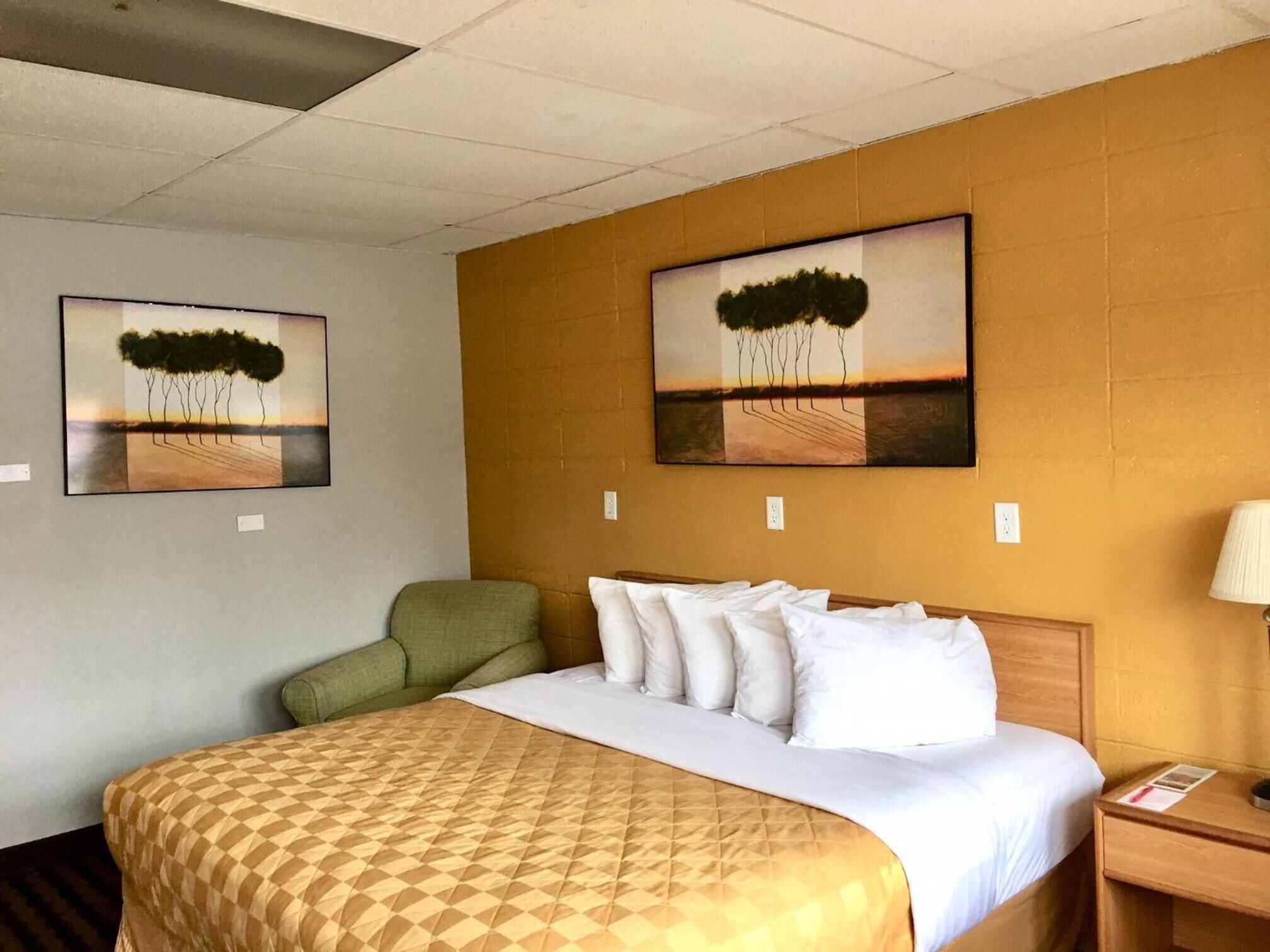 A hotel room with a large bed, a green armchair, two paintings on the wall, and a nightstand with a lamp.