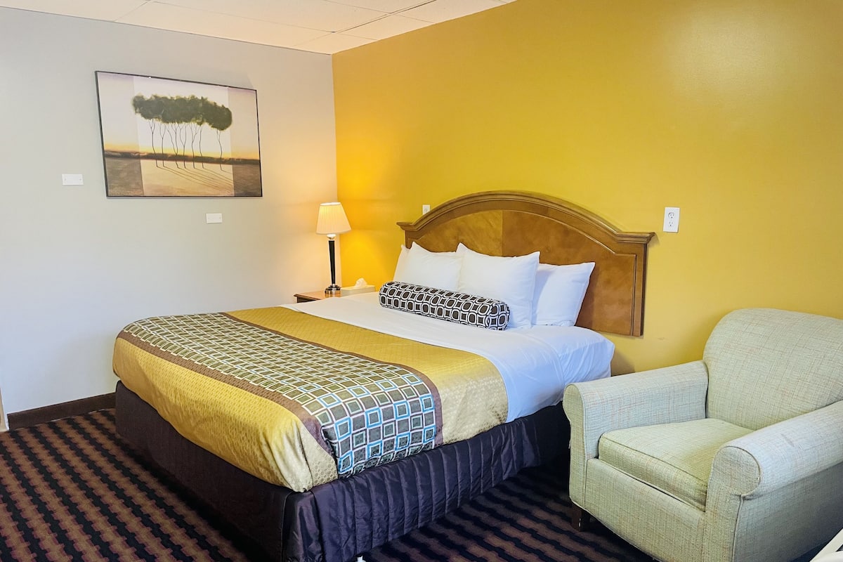 A hotel room with a bed, a chair, a lamp, and a painting on the wall.