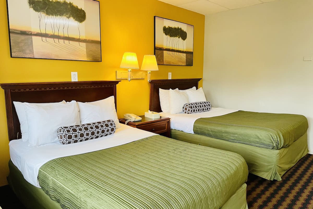 A hotel room with two beds, each with green bedding and white pillows. There are two wall-mounted lamps on the nightstands.