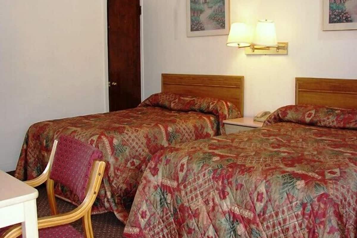 A hotel room with two beds, a wooden headboard, a chair, a desk, and a nightstand with a lamp.
