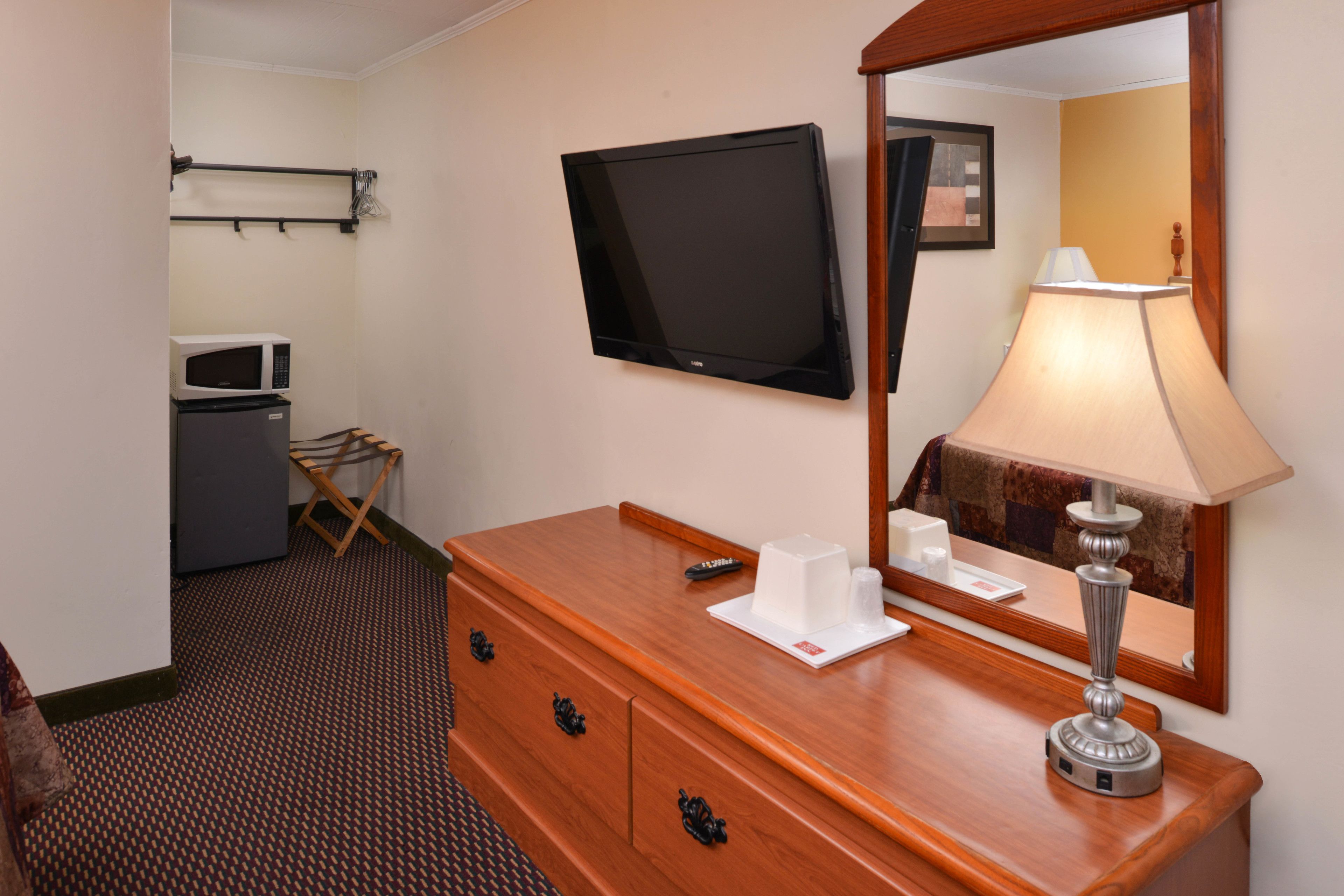 A hotel room with a wooden dresser, a flat-screen TV mounted on the wall, a lamp, a microwave, and a small table with a remote control.