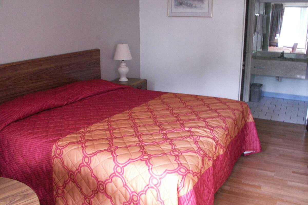 A bedroom with a wooden headboard, a bed with a red and beige quilt, a bedside table with a lamp, and a bathroom visible through an open door.