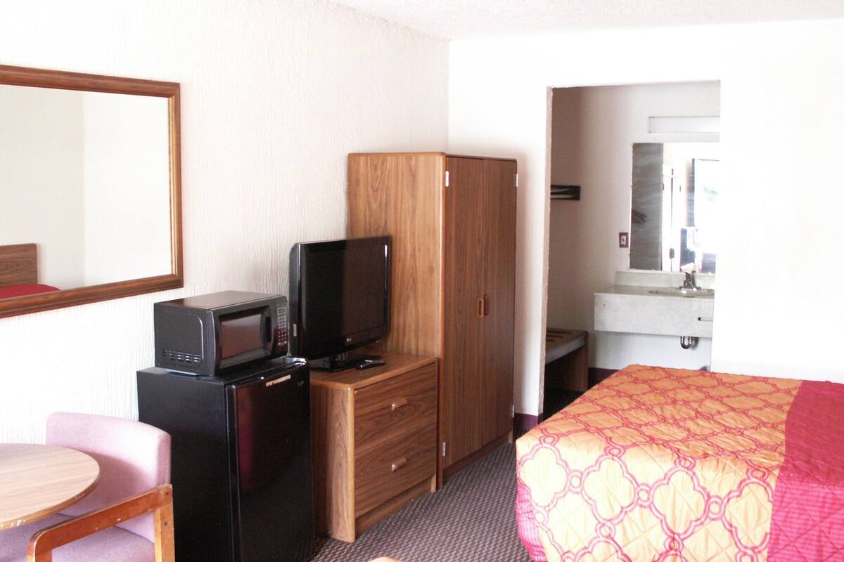 A hotel room with a bed, a wooden wardrobe, a desk with a microwave and a television, a chair, and a bathroom with a sink and mirror.