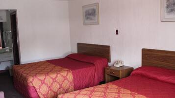A hotel room with two beds, each with a wooden headboard and red bedding. There is a bedside table with a telephone and a small drawer.