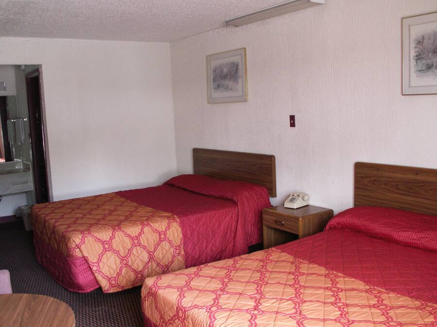 A hotel room with two beds, each with a wooden headboard and red bedding. There is a bedside table with a telephone and a small drawer.