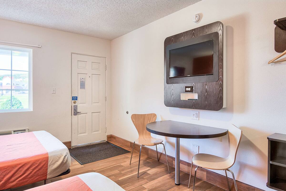 A room with two beds, a TV mounted on the wall, a round table with chairs, and a door.
