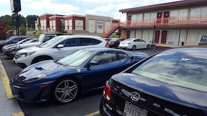 A parking lot with several luxury cars, including a blue Ferrari and a black Lexus. In the background, there is a building with a sign that reads "IMAX" and another sign for "CineBowl & Grill." - Red Carpet Inn Blacksburg (Blacksburg)