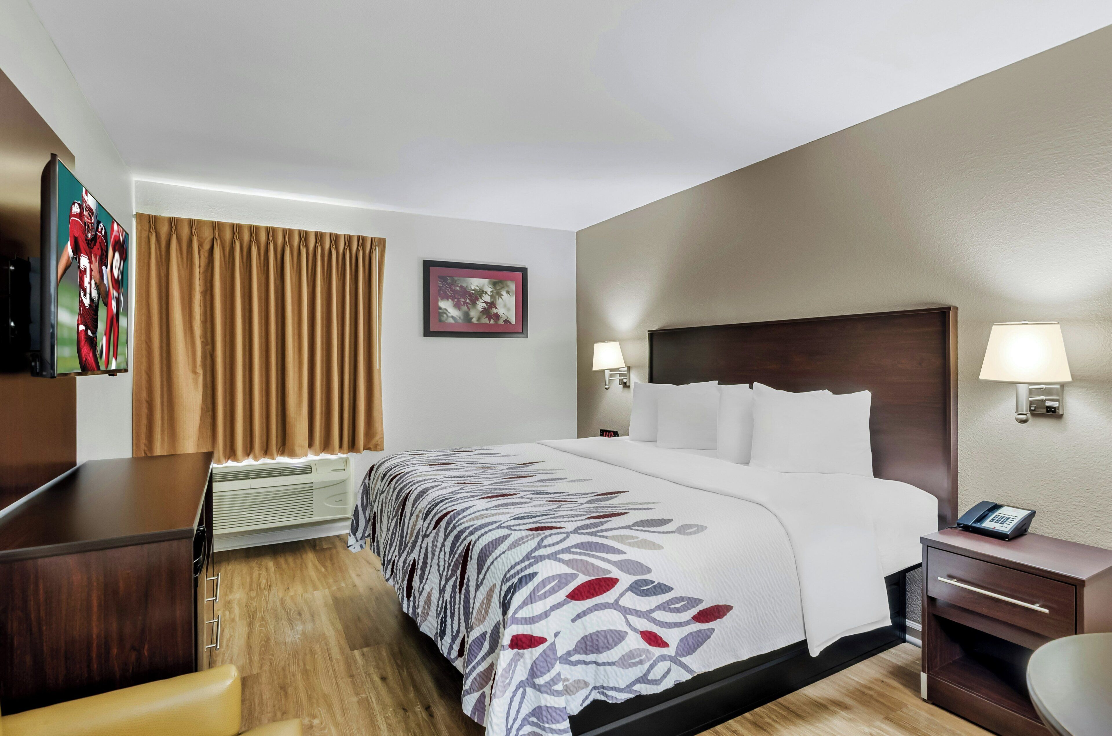 A hotel room with a bed, bedside lamps, a desk, a chair, a TV, and a framed picture on the wall.