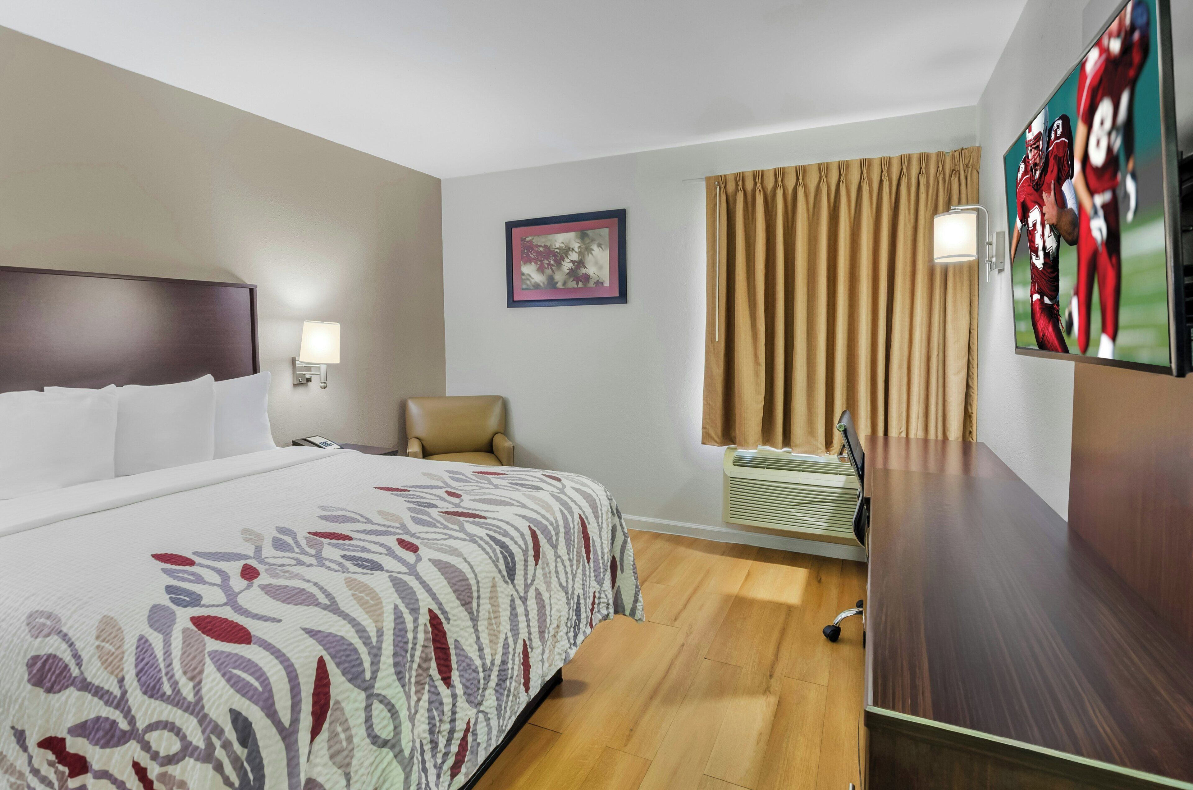 Habitación de hotel con cama, una silla, escritorio, televisor, un cuadro en la pared y ventana con cortinas.