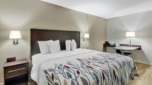 A hotel room with a bed, bedside lamps, a nightstand with a digital clock, a small table with a chair, and a wall-mounted shelf.
