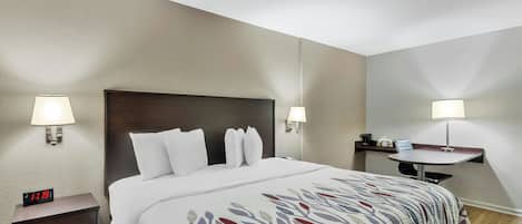 A hotel room with a bed, bedside lamps, a nightstand with a digital clock, a small table with a chair, and a wall-mounted shelf.