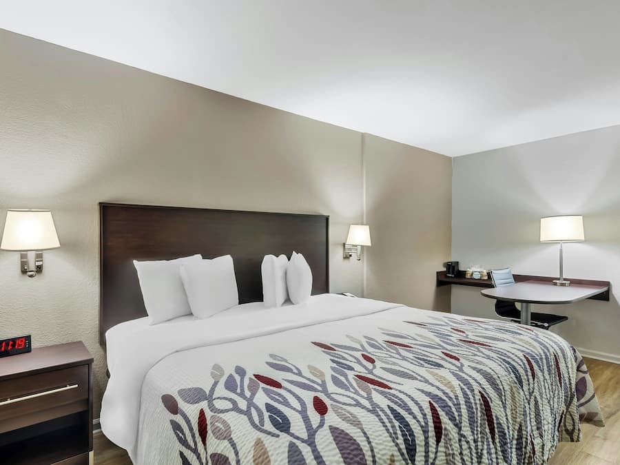 Ein Hotelzimmer mit Bett, Nachttischlampen, einem Nachttisch mit digitaler Uhr, einem kleinen Tisch mit Stuhl und einem an der Wand befestigten Regal.