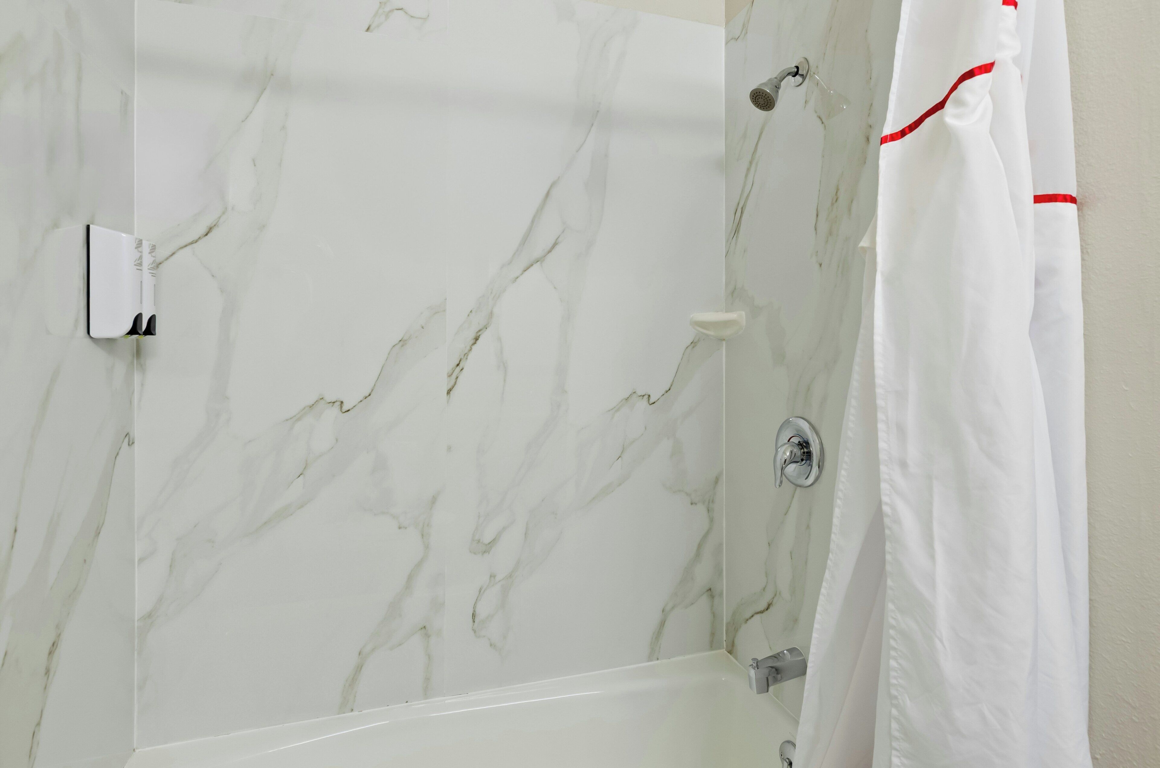 Une salle de bain avec un mur en marbre, une baignoire blanche et un rideau de douche blanc orné d’une bordure rouge.