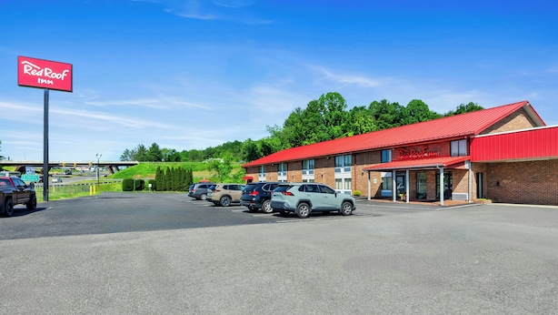 Un hôtel Red Roof Inn avec un toit rouge et une façade en briques, un parking avec plusieurs voitures et un ciel bleu dégagé.