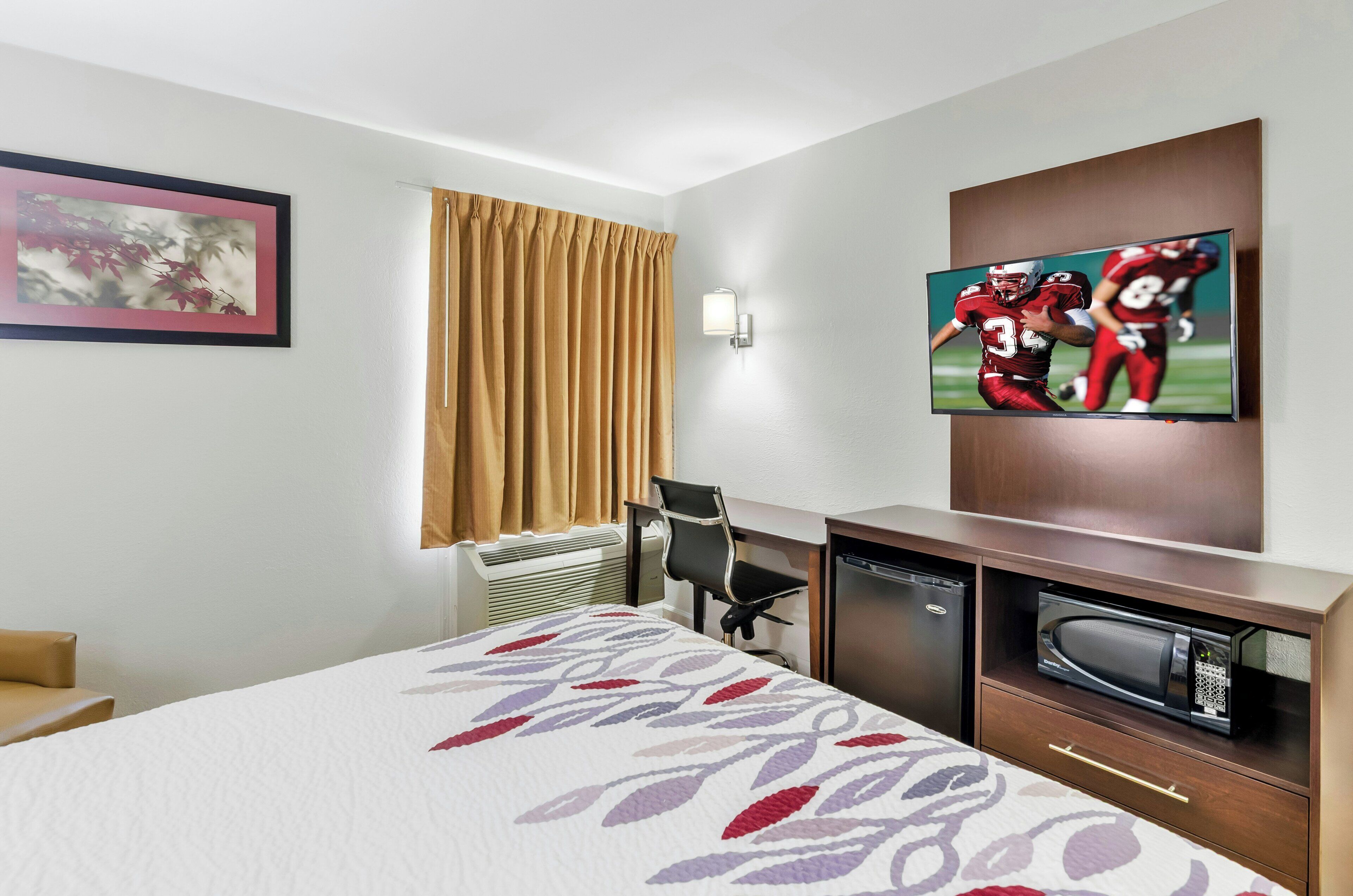 A hotel room with a bed, a desk with a microwave, a chair, a TV on the wall, and a window with curtains.