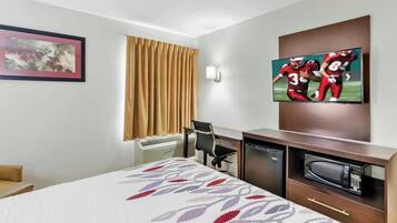 A hotel room with a bed, a desk with a microwave, a chair, a TV on the wall, and a window with curtains.