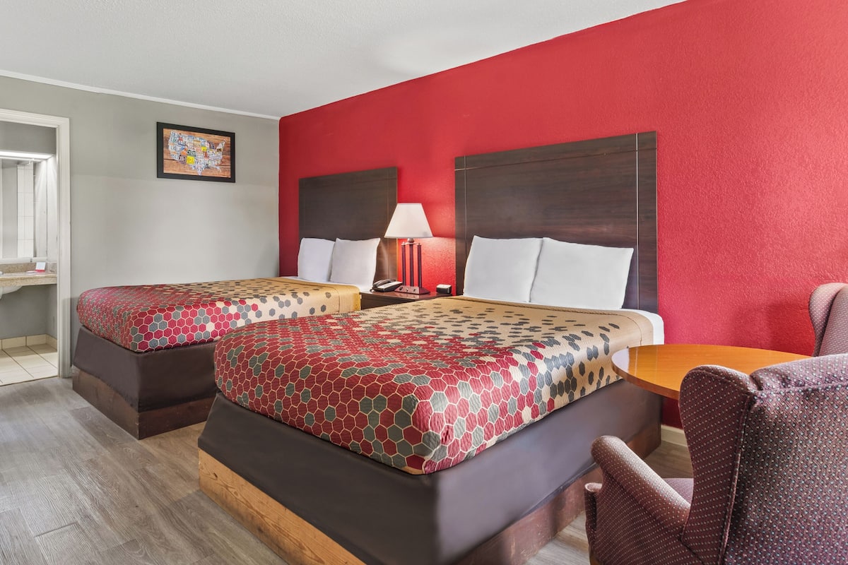 A hotel room with two beds, a wooden nightstand, a red wall, and a small table.
