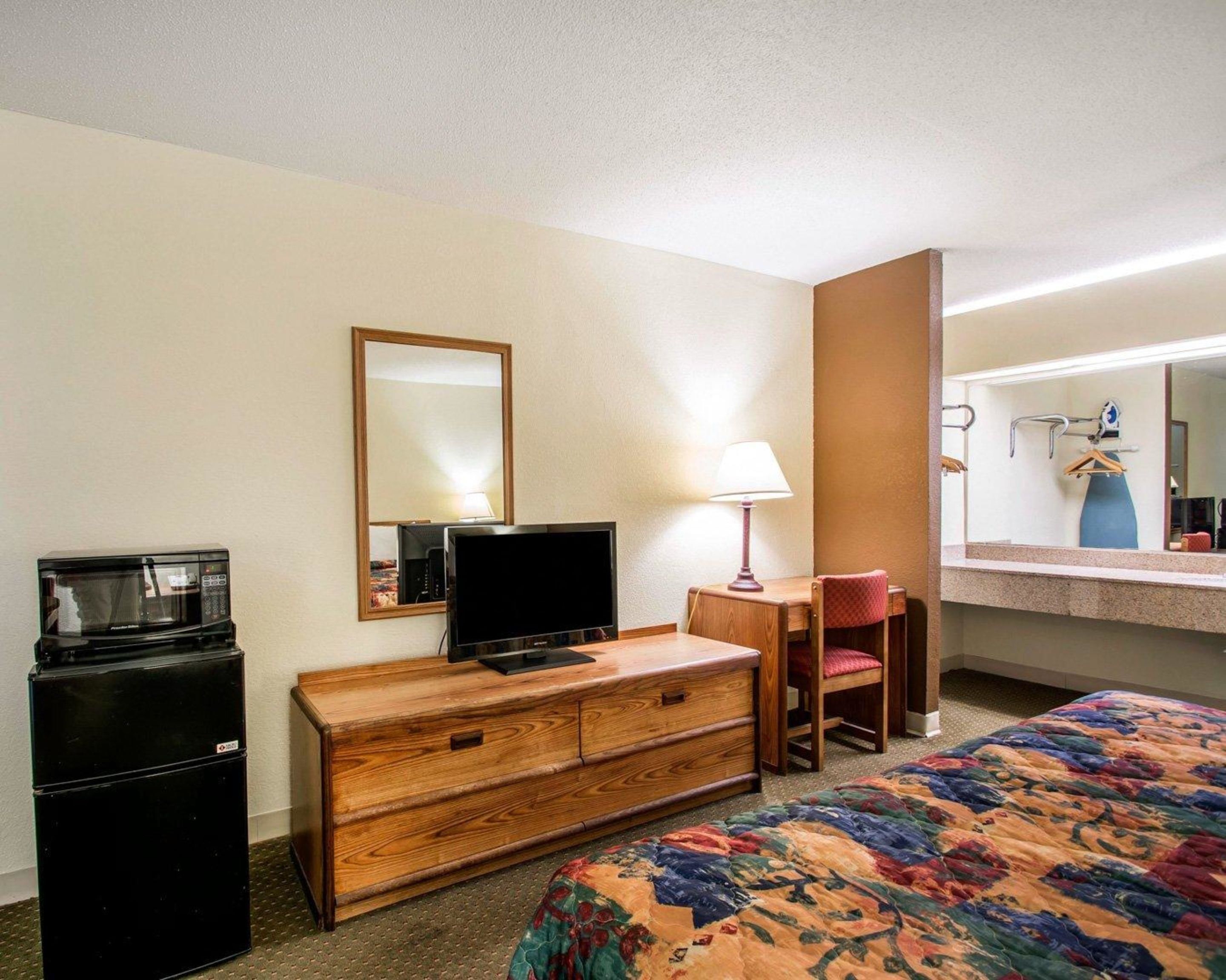 A hotel room with a bed, a wooden dresser, a television, a microwave, a desk with a chair, and a mirror.