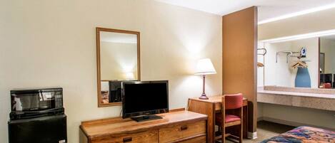 A hotel room with a bed, a wooden dresser, a television, a microwave, a desk with a chair, and a mirror.