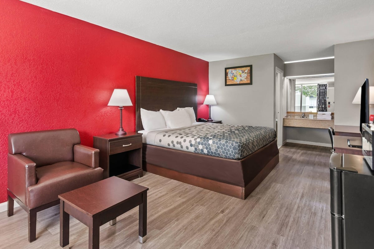 A hotel room with a bed, a chair, a table, a nightstand, a lamp, a TV, and a kitchen area with a refrigerator.