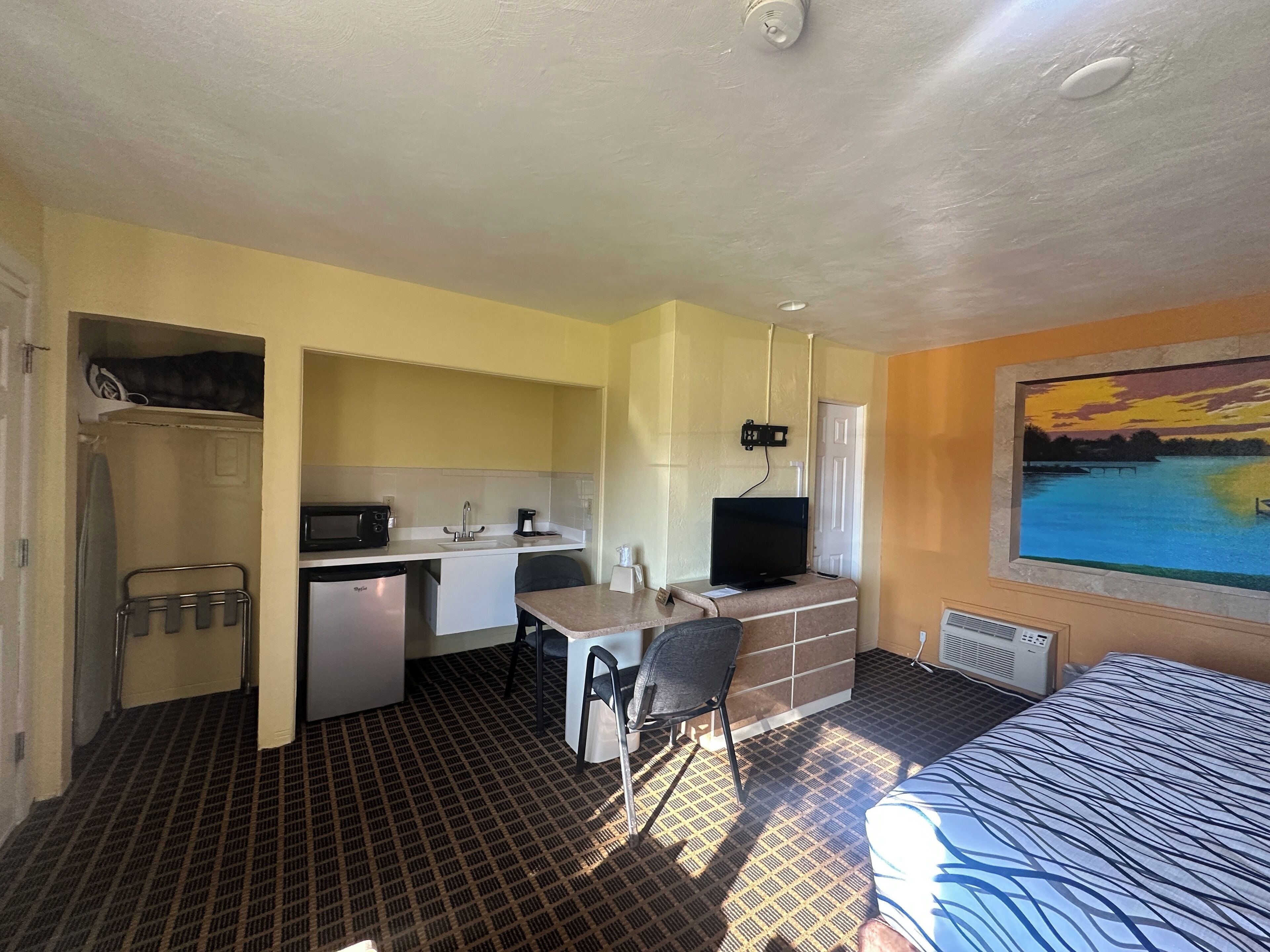 A compact hotel room with a kitchenette, a bed, a desk with a chair, and a painting of a sunset over a body of water.